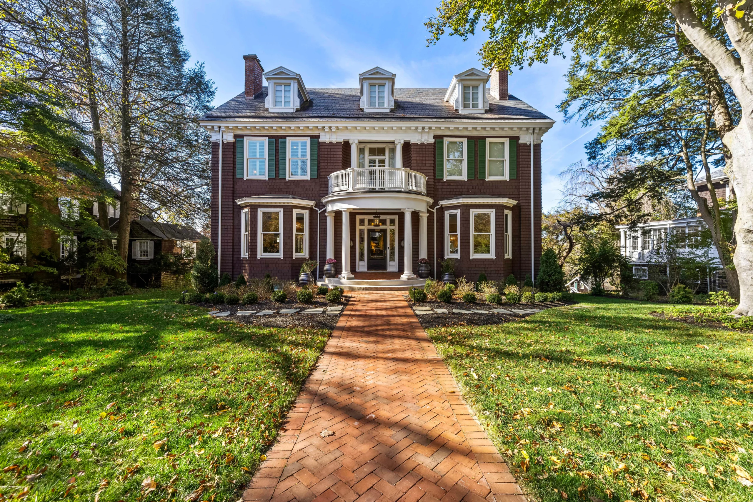 Front view of a red brick mansion with white trim, green shutters, and a curved balcony over the entrance, brick walkway leading up