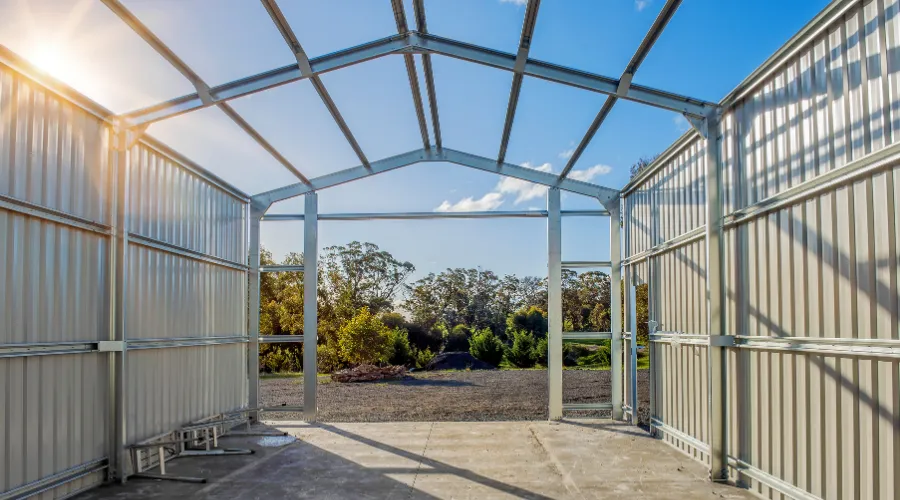 Metal framework of a small greenhouse with clear panels, sunlight streaming in from the left and a garden visible outside through the open doorway.
