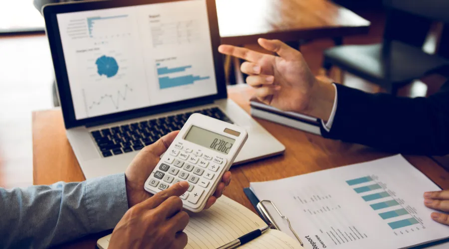 Two professionals discuss financial data: one uses a calculator while the other points at a laptop displaying charts and graphs on a wooden desk.